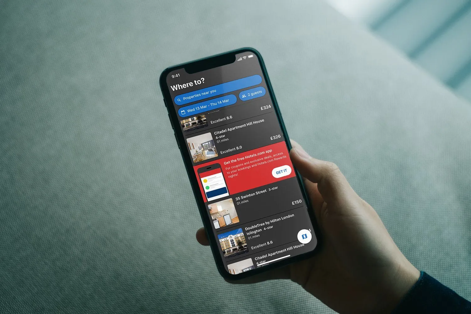 A hand holds a phone. The results list is dark, with a red banner for the free app, and hotels including Citadel Apartment Hill House and the DoubleTree by Hilton.