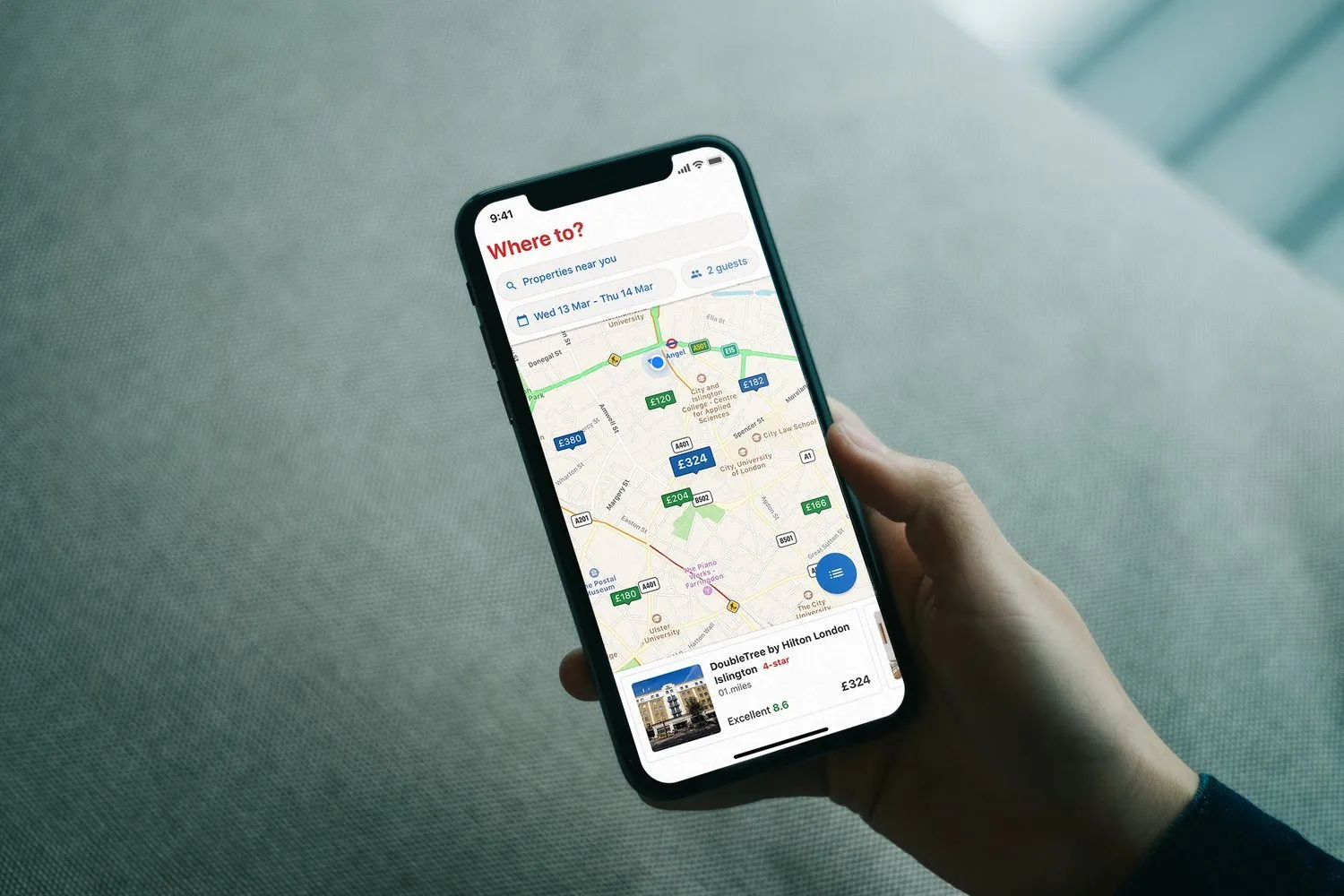 A hand holds a phone over grey fabric. The Hotels.com map is light, with price pins, and a card for the DoubleTree by Hilton London Islington at £324.