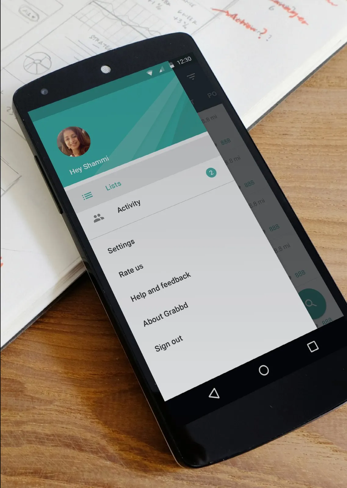An Android phone on a notebook. The Grabbd menu is open, greeting Shammi, with Lists, Activity, Settings, and Sign out.